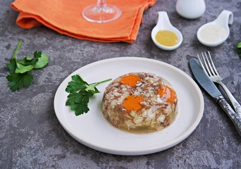 Cold appetizer, portioned meat jelly of pork, beef and chicken on a white clay plate on a gray concrete background. Holiday snacks for New Year. Russian cuisine. Selective focus.