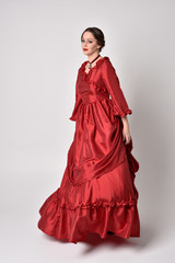 full length portrait of a brunette girl wearing a red silk victorian gown. Standing in side profile...