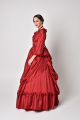 full length portrait of a brunette girl wearing a red silk victorian gown. Standing in side profile...