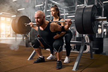 Strong brutal male powerlifter doing squats using heavy barbell, young active female trainer...