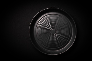 Closeup empty round black ceramic plate with pattern of circles on dark background with copy space. Overhead view. Concept modern shooting menu from chef, restaurant advert, tableware catalogs