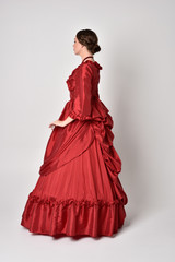 full length portrait of a brunette girl wearing a red silk victorian gown. Standing in side profile...