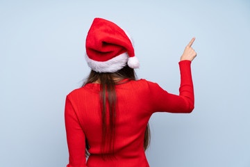 Girl with christmas hat over isolated blue background pointing back with the index finger