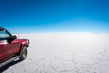 Fototapeta na wymiar A 4×4 driving tour travels across the vast, patterned Uyuni salt landscape towards the distant mountains on a clear blue sky day.