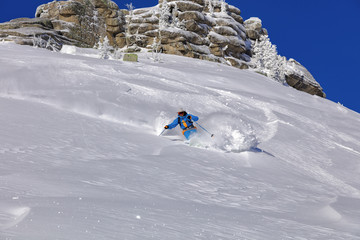 A skier on the slope. Out-of-piste skiing. Good winter day, ski season. professional rider in a bright blue and orange ski suit and hat on a background of blue sky and beautiful rocks