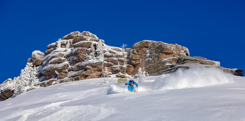 A skier on the slope. Out-of-piste skiing. Good winter day, ski season. professional rider in a bright blue and orange ski suit and hat on a background of blue sky and beautiful rocks