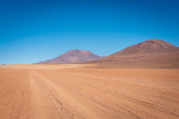 Fototapeta na wymiar Dirt tracks continue through the orange soil of the Siloli Desert travelling past countless volcanoes into the endless horizon on a clear blue sky day.