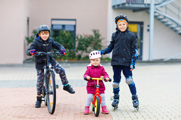 Two kids boys and little toddler girl playing together outdoors. Brothers in safety clothes skating...