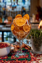 Glass full of dried sliced oranges, at a bar with rose petals sprinkled around for seasonal decoration. Bottles on shelves behind, out of focus.