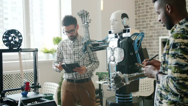 Cheerful guys scientists are testing cool robot in office with tablet, 3d printer is working in background. Modern technology and contemporary business concept.