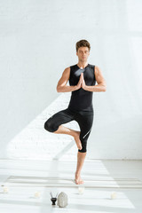 Fototapeta premium handsome young man practicing yoga in tree pose near decorative buddha head, aromatic sticks and candles