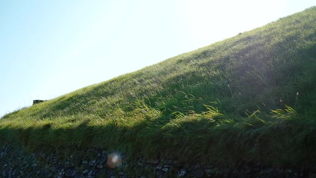 medium close sidewards pan and tilt up handheld shot of green grass-covered rooftop of the Newgrange Passage Grave against blue sky. Shadows on the stone walls and into the sun with beautiful flare.