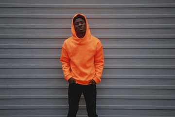 African American man wearing blank orange hoodie standing against grey wall - obrazy, fototapety, plakaty