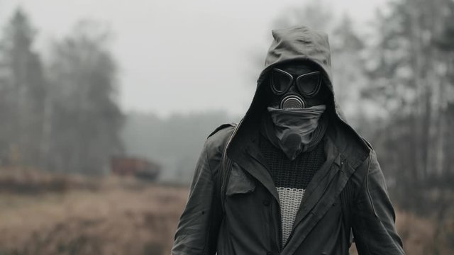 Portrait of man in gas mask standing by abandoned carriages at deserted railway station. He looks into camera turns out and go away. Armageddon concept, male wanderer survivor after nuclear disaster