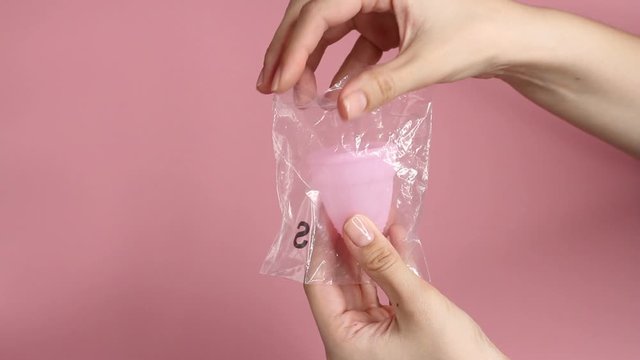 Close up of a young woman hands take out reusable pink silicone menstrual cup of the bag on pink background. Zero waste menstruation hygiene concept.