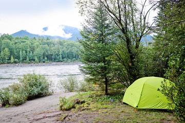 Tourist tent among bushes and trees on the banks of a mountain river. Bright tourist equipment.