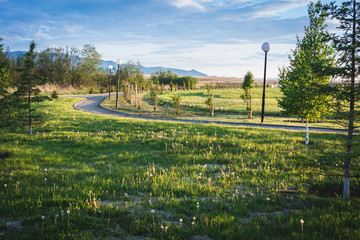 Winding alley with lanterns near the green grass for walk goes into the distance to the large mountains against the blue sky.