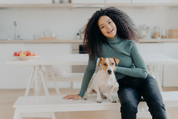 Female pet owner expresses love to dog, dressed in casual turtleneck and jeans, sits at white bench...