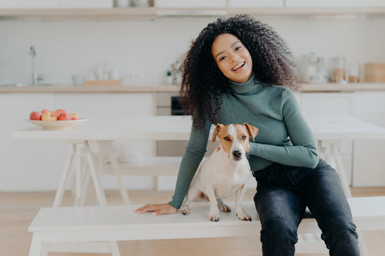 Female Pet Owner Expresses Love To Dog, Dressed In Casual Turtleneck And Jeans, Sits At White Bench Against Cozy Kitchen Interior, Spends Free Time At Home, Smiles Broadly From Happy Enotions