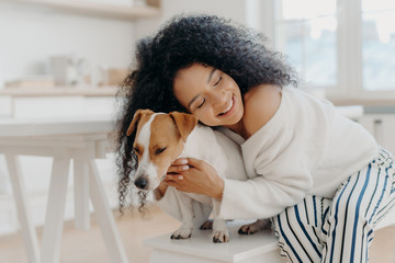 Lovely young curly African American woman embraces beloved pedigree dog with love, has gentle...