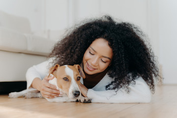 Relaxed Afro woman with crisp dark hair lies on floor, plays with cute puppy, has fun with jack...
