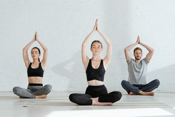 young men and woman practicing yoga in half lotus pose with raised prayer hands