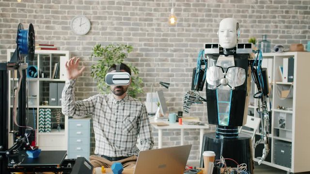 Man wearing ar glasses is moving hand while smart robot is repeating movements in office, 3d printer is visible. People, modern technology and work concept.