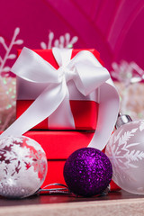 A small red gift box tied with white ribbon and violet and mat white Christmas balls with ornament. Cozy holiday home decorations