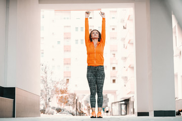 Good-looking fit healthy smiling caucasian woman in sportswear and with curly hair doing jumping jacks in passage on sunny day.