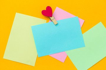 blank multicolored paper notes with cloth pin decorated with red heart on a vibrant yellow background