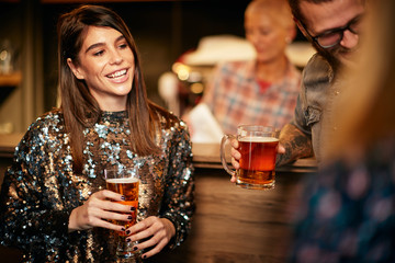 Beautiful caucasian smiling stylish woman  holding beer and chatting with friends. In background is bar counter. Pub interior.