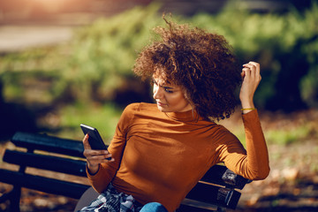 Serious mixed race young woman with curly hair and in turtleneck sitting in park on bench and using smart phone for reading or writing message.