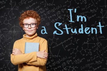 Cute student boy with ginger hair holding a book on chalkboard background