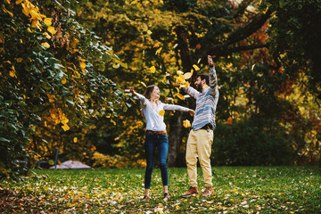 Naklejka na ściany i meble Playful caucasian happy young couple standing in nature and throwing leaves in air. Autumn time.