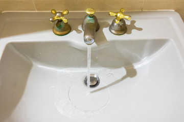 Water flows from an old metal faucet into a white ceramic sink. On the mixer parts there are...