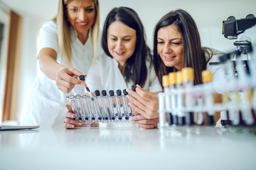 Three dedicated beautiful smiling caucasian female lab assistants holding and looking at test tubes...