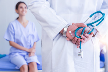 Doctor hold stethoscope behind back in patient room at hospital or clinic with smiley patient face background. Patient waiting doctor on hospital bed for health checkup. Health care concept. She happy