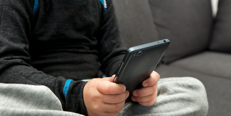 The little kid is sitting alone on the sofa and looking in his phone