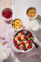 Bowl with oatmeal, corn flakes, raspberry and a glass of milk on a white wooden background