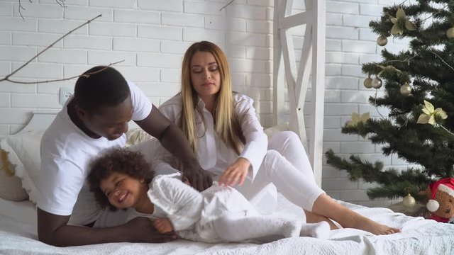 Young happy multi-ethnic family of mother, father and little daughter dressed in white at background of Christmas and New Year tree in home. Multi-racial family, Xmas concept