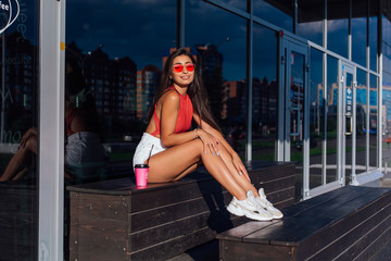 Stylish happy young brunette woman wearing white shorts and sneakers holding pink cup of coffee to go sitting next to coffee shop.