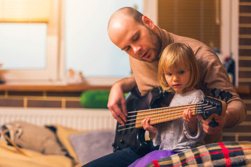 Cute little girl and her father are playing bass guitar and smiling while sitting on couch at home. Spending time together, learning to play the guitar