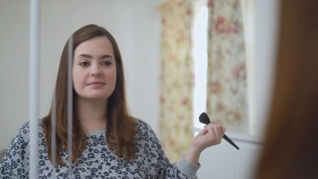 Woman applying blush with a brush in front of the mirror. Caucasian female using beauty cosmetics to improve herself ready to work in bedroom at home.