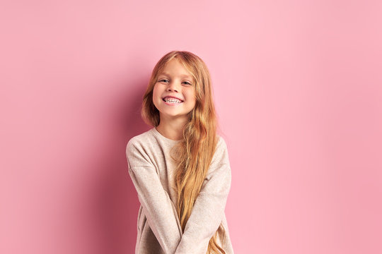 Portrait of positive cheerful girl cutely smiling at camera, girl with long golden hair in white blouse. Pink background