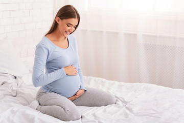 Pregnant Lady Touching Belly Smiling Sitting On Bed At Home