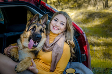 A happy Caucasian girl lies in trunk of a car with a German shepherd dog breed and smiles, a...