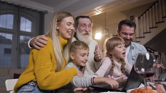 Charming family of adults and children sitting at the table and saying goodbay with their computer interlocutors during video call