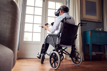 Mature Disabled Man In Wheelchair At Home Wearing Virtual Reality Headset Holding Gaming Controllers