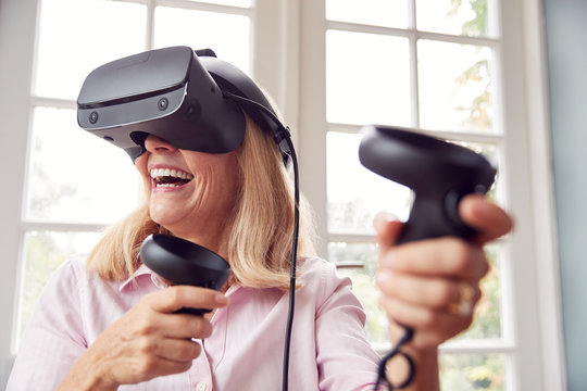 Mature Disabled Woman In Wheelchair At Home Wearing Virtual Reality Headset With Gaming Controllers
