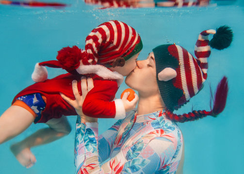 Caucasian woman (mother) kissing her child (infant) underwater in new year and christmas elf suits in the swimming pool